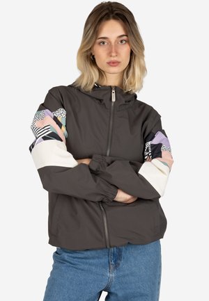 Junge Frau mit blonden Haaren, die eine dunkle Jacke mit Reißverschluss und mehrfarbigen geometrischen Mustern auf den Ärmeln sowie blaue Jeans trägt, die Arme verschränkt.