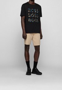Svart kortärmad t-shirt med flerfärgad "BOSS" logotyp, tillsammans med beige shorts. Svarta skor och strumpor fulländar outfiten.