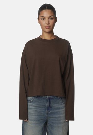 Donna con capelli scuri legati dietro che indossa una maglia marrone a maniche lunghe e jeans azzurri su uno sfondo uniforme.