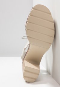 Beige sko sula med räfflad yta, försedd med små metallnitar längs kanten. Slät yta och rundad form.
