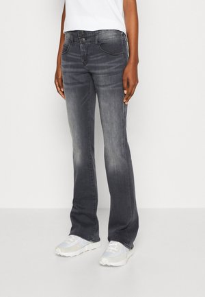 Person iført falmede sorte bootcut jeans og hvide sneakers, stående mod en ensfarvet hvid baggrund.