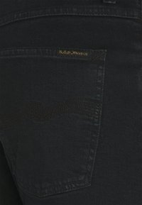 Mörka denimjeans med en bakficka som har ett subtilt vågmönster och en guldlogotagslapp som säger "Nudie Jeans Co." på fickans söm.