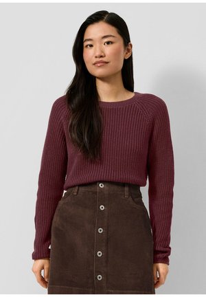 Femme aux longs cheveux foncés porte un pull en maille bordeaux et une jupe boutonnée en velours côtelé marron sur un fond gris uni.