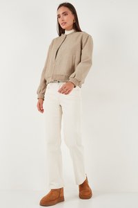 Kvinde iført beige knap-op jakke, hvide højttaljede bukser og brune ankelstøvler, stående mod en ensfarvet lys baggrund.