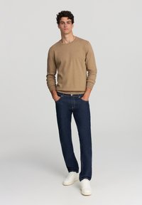 Maglione beige a girocollo, jeans blu scuro e sneakers bianche. Il modello sta in piedi con le mani nelle tasche su uno sfondo neutro.