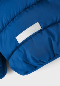 Modrá nafukovací bunda s hladkým nylonovým materiálem, horizontálním prošíváním a kontrastní bílou logovou nášivkou.