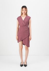 Kobieta stoi na białym tle, ubrana w bezrękawą, teksturowaną sukienkę w odcieniu mauve z warstwowym krojem oraz czarne, spiczaste szpilki.