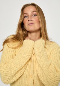 Dzeltena adīta kardigāna ar biezām tekstūru, ribotām piedurknēm un lielām pogām, kas izceļas ar vaļīgu piegriezumu un patīkamu izskatu.