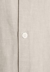 Gros plan sur un tissu beige clair avec un bouton blanc à quatre trous cousu à côté d'une couture verticale.