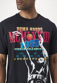Zwarte katoenen t-shirt met gedurfde gekleurde graphics, waaronder "PUMA HOOPS," een basketbal en een silhouet van een speler. Opvallende datum en steraccenten.