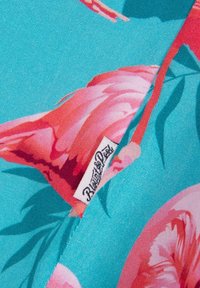 Un tissu turquoise vif orné de grands motifs floraux roses. Une étiquette avec le texte "Blue Sky" est visible, mettant en avant les détails de la marque.
