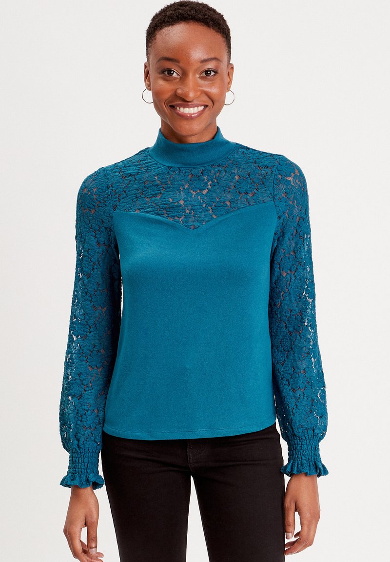 Top a maglia turchese con maniche lunghe in pizzo trasparente e scollatura alta, caratterizzato da un corpino aderente e polsini arricciati.