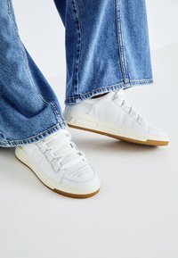 Weiße Leder-Sneaker mit perforierten Details, flachen weißen Schnürsenkeln und einer Gummisohle, kombiniert mit weit geschnittenen blauen Jeans.