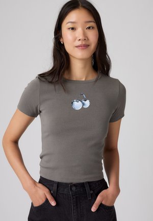Giovane donna con lunghi capelli scuri che indossa una maglietta grigia aderente con una grafica di due ciliegie blu e pantaloni neri, mani nelle tasche.