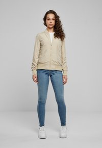 Blouson aviateur beige avec une fermeture éclair à l'avant, des poignets et une ceinture côtelés. Porté sur une chemise blanche avec un jean skinny bleu et des baskets blanches.