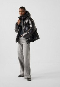 Chaqueta de plumón negra con un acabado brillante, capucha oversized y cierre de cremallera, combinada con jeans de pierna ancha grises y zapatos negros con tachuelas.