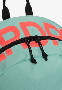 Mochila verde menta con tiradores de cremallera negros y texto parcial en rojo intenso cerca de la cremallera, mostrando una textura de tela detallada y costuras.