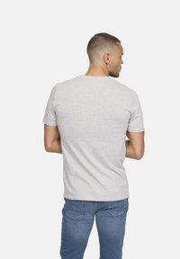 Homme aux cheveux courts portant un t-shirt gris clair et un jean bleu, se tenant légèrement tourné vers la droite devant un fond blanc uni.