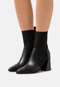 Botas de tobillo negras con tacón y puntera puntiaguda, con un exterior de cuero y un caña de tela acanalada, combinando materiales lisos y texturizados.