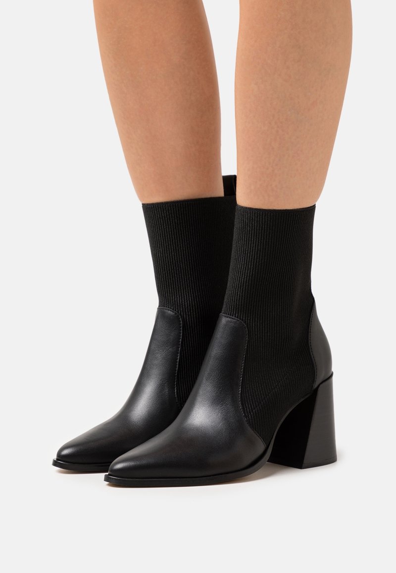 Botas de tobillo negras con tacón y puntera puntiaguda, con un exterior de cuero y un caña de tela acanalada, combinando materiales lisos y texturizados.