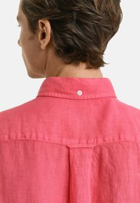 Camicia di lino rosa vista dal retro, con colletto con bottoni e un dettaglio a piega vicino alle spalle. Texture liscia, design informale.