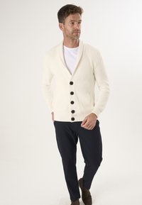 Valge kudumiskardigan, millel on šaalikaelus, viie suure musta nupuga, kantud valge T-särgi ja tumesiniste pükstega.