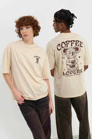 Beige oversized t-shirt featuring a koala graphic with a coffee cup on the back and smaller text on the front. Worn with dark pants.