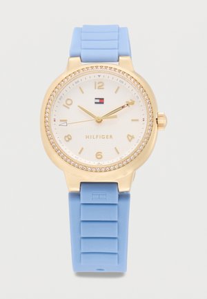 Goldfarbene runde Uhr mit Kristall-Lünette, weißem Zifferblatt, goldenen Zahlen und Zeigern, blauem Silikonarmband und Tommy Hilfiger-Logo auf Zifferblatt und Armband.