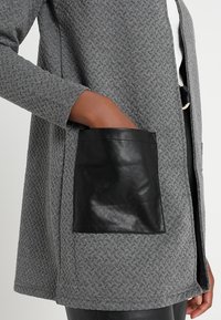 Lähikuva henkilöstä, joka pitää yllä tekstuurista harmaata takkia, jossa on suuri mustasta nahasta valmistettu tasku, ja hänen kätensä on taskussa.