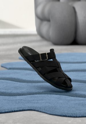 Black leather slip-on sandal with buckle resting on textured blue rug, blurred rounded furniture in neutral-toned room background.
