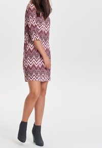 Vestido con un patrón en zigzag rosa y morado, hasta la rodilla, con mangas de tres cuartos. Combinado con botines de tacón grises.