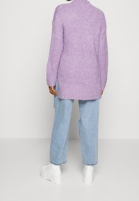 Lavendelstickad tröja med hög hals, långa ärmar och sidokilar, i kombination med ljusblå baggy jeans och vita plattformsskor.