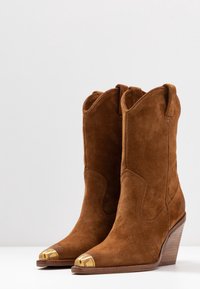 Botas occidentales de ante marrón con puntera afilada, punta metálica dorada y tacón de cuña apilada. Presentan costuras contrastadas y lengüetas de agarre.