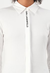 White long-sleeved shirt with a classic collar, featuring a front closure and a vertical black logo text "KARL LAGERFELD" on the placket.