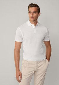 Polo shirt bianco a maniche corte con colletto e una leggera texture. Presenta un piccolo logo ricamato sul petto. Abbinato a pantaloni beige chiaro.