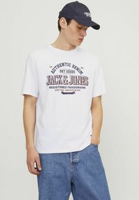 Hvid bomulds t-shirt med et trykt logo design, der læser "JACK & JONES" i marineblå og burgunder. Bæres med blå denim jeans.