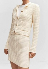 Cardigan tricoté couleur crème avec un motif gaufré texturé, doté de deux poches avant et de trois boutons en bois. Associé à une jupe assortie.