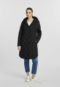 Czarny, pikowany długi płaszcz z kapturem, z zapięciem na guziki z przodu i kieszeniami po bokach. Nosi się go na niebieskie dżinsy i białe trampki.