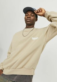 Beige sweatshirt with a crew neckline, ribbed cuffs, and hem. Features a white logo on the left chest. Smooth texture. Paired with a dark shirt underneath.