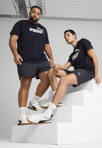 Námořnické trička Puma s bílým logem, odpovídající šortky a bílé boty na modelu. Jeden model sedí na bílých schodech a předvádí design obuvi.