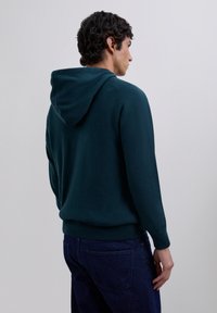 Man som vänder bort med ryggen, iförd en mörkgrön huvtröja och mörkblå jeans mot en enfärgad ljusgrå bakgrund.