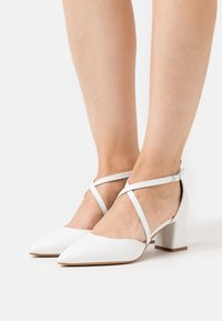 Bílé kožené boty na blokovém podpatku s špičatými špičkami, zdobené křížovou pružnou páskou kolem kotníku a hladkým matným povrchem. Klasický design.