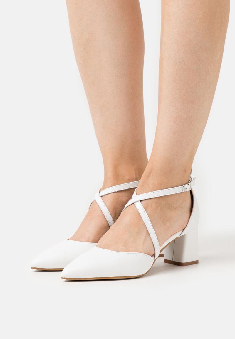 Bílé kožené boty na blokovém podpatku s špičatými špičkami, zdobené křížovou pružnou páskou kolem kotníku a hladkým matným povrchem. Klasický design.