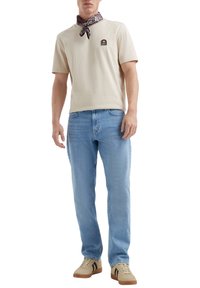 T-shirt en coton beige à manches courtes, patch logo, jean denim bleu clair, baskets beiges avec rayures noires et bandana marron à motifs.