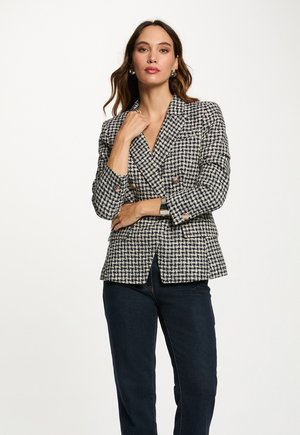 Vrouw in een zwart-wit pied-de-poule blazer met gouden knopen, donkere jeans, zilveren oorbellen en een zilveren armband, die zelfverzekerd poseert.