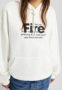 Sudadera blanca con capucha y un bolsillo delantero. El diseño presenta texto negro, incluyendo la palabra "Fuego", en diferentes tamaños y estilos.