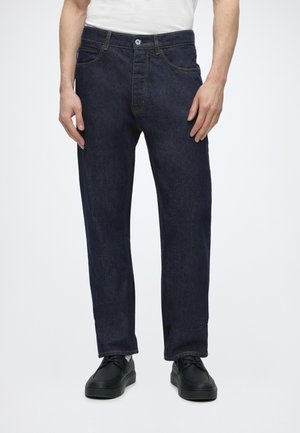Man die donkerblauwe rechte jeans, zwarte schoenen en een wit overhemd draagt, staand tegen een effen witte achtergrond.
