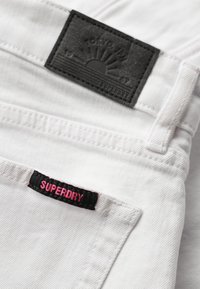 Jean de mezclilla blanca con una etiqueta negra texturizada que presenta un sol y "SUPERDRY" grabado. Una etiqueta rosa con "SUPERDRY" cosida en el bolsillo.