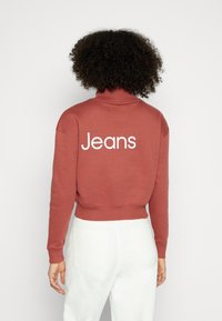 Sudadera recortada de color óxido con cuello alto y texto blanco "Jeans" en la parte posterior. El material parece suave y tiene un diseño casual.