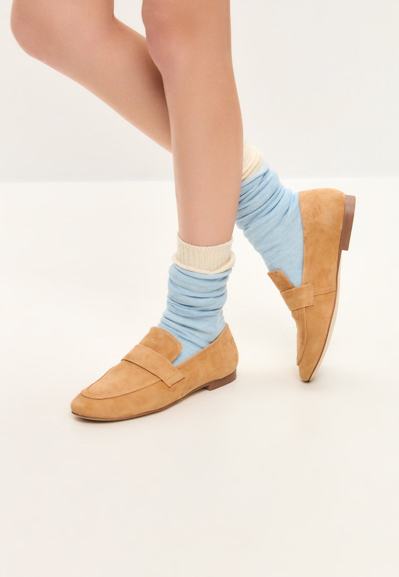 Mocassini in suede marrone chiaro con suola piatta e dettaglio a fascia rettangolare, abbinati a calzini morbidi blu e crema. Texture liscia e design classico.
